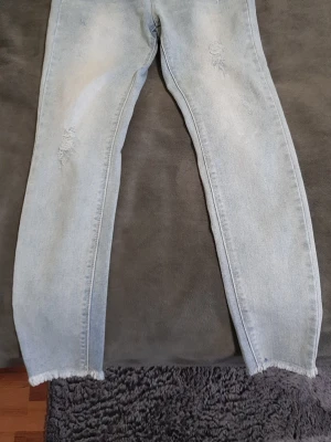 Ljusblå  jeans med slitningar - Fina jeans. Säljer ett par ljusblå jeans med och slitna detaljer på låren. Jeansen har råa, fransiga benslut och är tillverkade i klassisk denim. Perfekta för en avslappnad och trendig look.