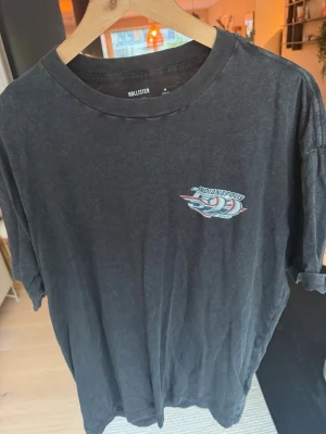 Svart boxy t-shirt från Hollister - Säljer en svart t-shirt från Hollister i boxy fit med tryck på bröstet som har racingtema och texten 'Indianapolis'. Tillverkad i 100% bomull och har en avslappnad passform med korta ärmar.