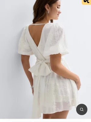 Vit omlottklänning med rosett - Söt vit kortklänning i omlottmodell med puffärmar och v-ringning. Klänningen har en stor rosett i ryggen och är tillverkad i ett lätt, blommigt mönstrat tyg som ger en romantisk känsla. Perfekt för sommarens alla tillfällen. Storlek M men jag som har normalt S kunna ha den då det går att dra åt i midjan! 