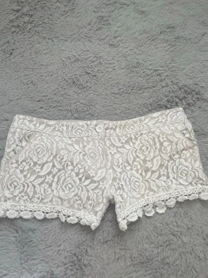 Vita spets-shorts med blommönster - Säljer ett par vita shorts med vackert blommigt spetsmönster och dekorativ volangkant längst ner. De har knapp och dragkedja framtill och är perfekta för sommarens alla tillfällen. Supersnygga att matcha med en enkel topp!