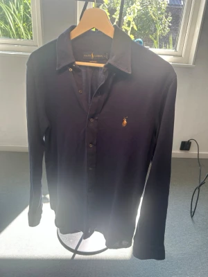 Mörkblå långärmad skjorta Polo Ralph Lauren - Snygg mörkblå långärmad skjorta från Polo Ralph Lauren med klassisk krage och knappar framtill. Skjortan har den ikoniska broderade loggan på bröstet och är tillverkad i mjukt bomullstyg. Perfekt för en stilren look.