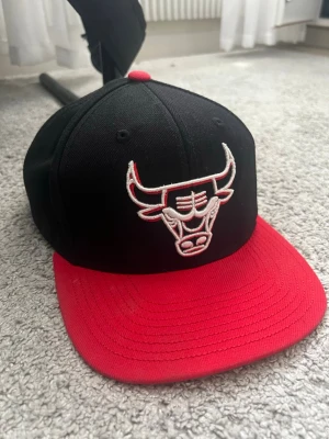 Svart och röd keps med tjurmotiv - Snygg keps i svart med röd skärm och broderad tjur på framsidan. Justerbar snapback i rött och vitt baktill. Perfekt accessoar för dig som gillar sportig streetstyle och vill sticka ut med en klassisk look.