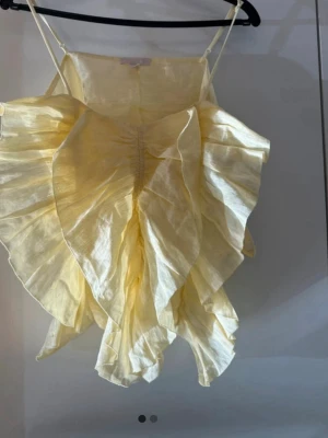 Gul volangtop från H&M - Säljer en ljusgul topp från H&M med tunna axelband och stora volanger framtill. Toppen har en luftig och flowy känsla, perfekt för sommardagar. Materialet ser ut att vara lätt och något glansigt, vilket ger en extra cool look.