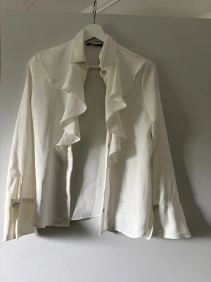 Vit blus med volang och pärlknappar - Stilren vit blus med långa ärmar och volangdetaljer längs kragen och framtill. Blusen har dekorativa pärlknappar vid halsen och ärmsluten. Tillverkad i satin, ett lätt tyg som ger ett elegant intryck. Perfekt för dig som gillar en chic och klassisk look.