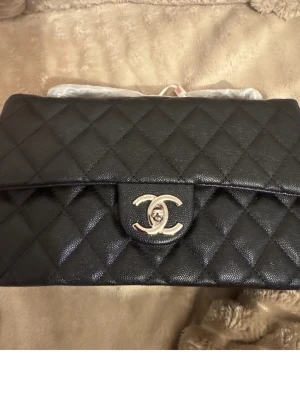 Svart quiltad handväska - Stilren svart handväska med quiltat mönster och rektangulär form. Väskan har en ficka på baksidan och ett lock framtill. Tillverkad i lambskin med struktur. Lång axelrems kedja. Perfekt för dig som gillar klassisk och trendig design.