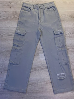 Grå cargobyxor med slitningar - Säljer ett par beige cargobyxor med raka ben och flera stora fickor på sidorna. Byxorna har slitna detaljer vid knäna och en avslappnad passform. Perfekta för en streetwear-look och passar till sneakers eller boots.