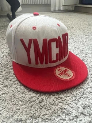 Vit och röd YMCMB snapback keps - Säljer en snygg snapback keps med YMCMB-brodyr i rött framtill. Kepsen är vit med röd skärm och röda detaljer. Klassisk platt skärm och justerbar baktill. Perfekt för dig som gillar streetwear och vill sticka ut.