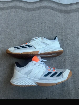 Vita Adidas sneakers med gummisula - Snygga vita Adidas sneakers med klassiska svarta ränder på sidan och detaljer i orange. Skorna har en slitstark gummisula och ovandel i mesh och syntetmaterial. Perfekta för dig som gillar sportig stil och vill ha bekväma skor till vardags.