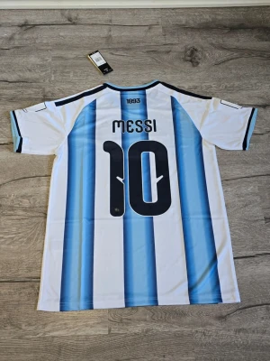 Argentina Messi matchtröja 2026 - Säljer en officiell Argentina matchtröja från Adidas med Messi och nummer 10 på ryggen. Tröjan har klassiska blå och vita ränder, svarta detaljer och guldiga loggor på bröstet. Tillverkad i lätt och ventilerande material, perfekt för fotboll eller träning.