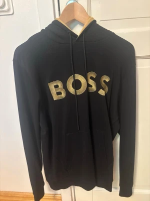 Svart hoodie från BOSS med guldtext - Snygg svart hoodie från BOSS med stor guldfärgad logga på bröstet. Hoodien har huva med dragsko och en klassisk magficka framtill. 