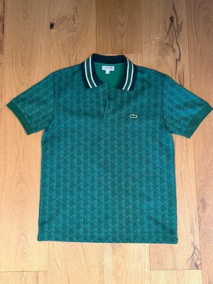 Grön pikétröja från Lacoste - Snygg grön pikétröja från Lacoste med diskret geometriskt mönster och klassisk krage med vita ränder. Tröjan har korta ärmar och den ikoniska krokodilloggan broderad på bröstet. Perfekt för en stilren och sportig look.