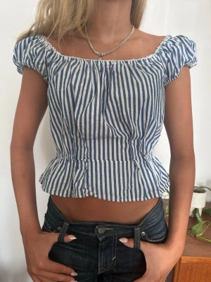 Blåvit randig croppad blus - Söt croppad blus med blå och vita vertikala ränder. Toppen har korta puffärmar, resår vid axlarna och en volang nedtill som ger en feminin siluett. Perfekt för varma dagar och enkel att styla med jeans eller kjol. Har en svag fläck på ryggen som knappt märks❣️