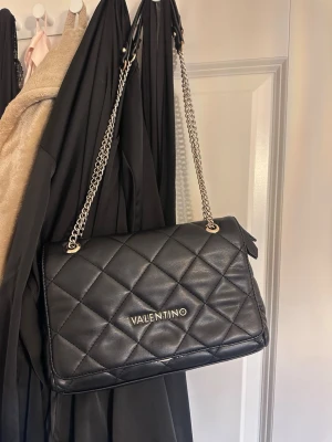 Svart quiltad handväska Valentino - Snygg svart quiltad handväska från Valentino med silverfärgad kedjerem och logga framtill. Väskan har ett stort huvudfack, innerficka med dragkedja och magnetlås. Tillverkad i läderimitation med elegant rutmönster.