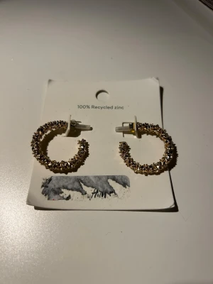 Guldiga creoler med pärldetaljer - Snygga creolörhängen i guldton med små pärldetaljer som ger ett glittrigt intryck. Tillverkade av 100% återvunnet zink. Perfekta för att lyfta din stil med en trendig och elegant touch. Oanvända.