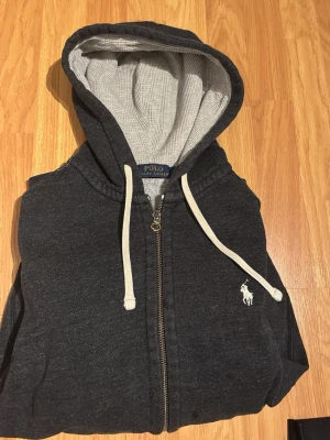 Mörkgrå hoodie från Polo Ralph Lauren - Mörkgrå zip-hoodie från Polo Ralph Lauren med vit broderad logga på bröstet. Hoodien har vit dragsko, fickor framtill och mjukt grått innerfoder i huvan. Perfekt för en avslappnad och stilren look.
