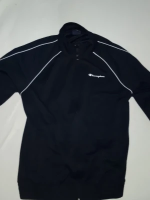 Svart half zip tröja Champion - Svart långärmad tröja från Champion med half zip och vita kontrastlinjer längs ärmar och axlar. Champion-logga på bröstet. Perfekt för en sportig look och passar till både träning och chill.