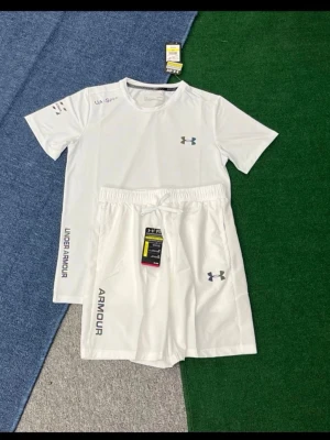Vitt träningsset från Under Armour - Snyggt vitt träningsset från Under Armour med t-shirt och shorts. Båda plaggen har logga och textdetaljer i grått. T-shirten har rund hals och korta ärmar, shortsen har resår i midjan och dragsko. Perfekt för gym eller löpning.