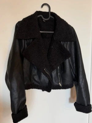 Svart teddyjacka med bred krage - Säljer en svart teddyjacka med bred, fluffig krage och muddar. Jackan har en croppad passform, dragkedja framtill och detaljer i mjukt teddy-material. Perfekt för dig som vill ha en trendig och mysig look.