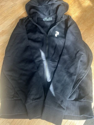 Svart hoodie från Peak Performance - Säljer en svart hoodie från Peak Performance med dragkedja framtill och huva. Hoodien har ribbade muddar vid ärmslut och nederkant samt en diskret logga på bröstet. Perfekt för en avslappnad och sportig stil.