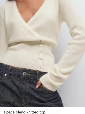 Krämvit omlottopp i alpaca-mix - Snygg krämvit stickad omlottopp i mjuk alpaca-blandning. Modellen har långa ärmar, djup v-ringning och dekorativa knappar i sidan. Perfekt att styla med jeans för en trendig look.