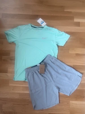 Nike träningsset mintgrön & grå - Snyggt träningsset från Nike med en mintgrön t-shirt och ljusgrå shorts. T-shirten har rund hals och diskret Nike-logga på bröstet. Shortsen är lätta och har elastisk midja samt liten Nike-logga. Perfekt för gymmet eller löprundan.
