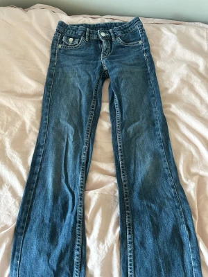 Blå bootcut jeans med fickdetaljer - Säljer ett par klassiska blå bootcut jeans med snygga fickdetaljer och kontrastsömmar. Jeansen har två framfickor, två bakfickor med lock och knapp samt låg till mellan hög midja. Perfekta för en avslappnad och trendig look.