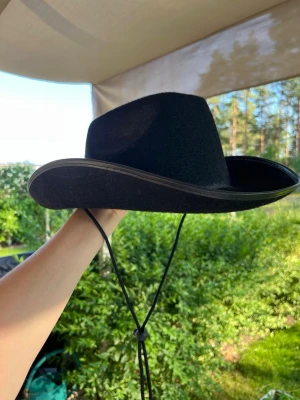 Svart cowboyhatt med snöre - Snygg svart cowboyhatt med ett justerbart hakband. Hatten har en klassisk westernstil. I bilden kan ljuset göra att den ser lite sotig ut men ser inte ut så i verkligheten. 