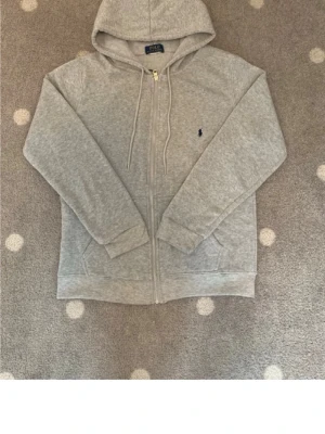 Grå zip hoodie från Polo Ralph Lauren - Snygg gråmelerad hoodie från Polo Ralph Lauren med dragkedja, huva och två fickor framtill. Klassisk blå broderad logga på bröstet. Tillverkad i mjukt sweatshirtmaterial som är perfekt för chill dagar. Storleks lappen har åkt av men jag beställde S.