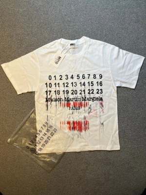 Maison Margiela vit t-shirt med tryck - Vit t-shirt från Maison Martin Margiela med ikoniskt siffertryck och text på bröstet. T-shirten har korta ärmar och en loose passform. Materialet är mjuk bomull och trycket har en cool, sliten effekt med röda och svarta detaljer.