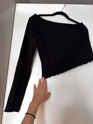 Svart oneshoulder topp med lång ärm - Trendig svart asymmetrisk oneshoulder topp med lång ärm. Toppen är lite croppad och har vågig kant längst ner för en extra snygg detalj. Perfekt att styla med höga jeans eller kjol. Storlek M men känns som en S då armen är ganska kort! 