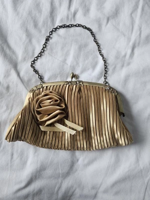✨ Superfin 90s vintage festväska i guld ✨ - Säljer en otroligt fin plisserad aftonväska i guldsatin från 90-talet. Den har en jättevacker rosdetalj på framsidan och stängs med en elegant metallbygel och kedja.Väskan är i superfint skick, inga fläckar eller skador alls! Perfekt till bal, bröllop, studenten eller festen.Fraktar gärna via Plicks säkra fraktsystem 📦✨