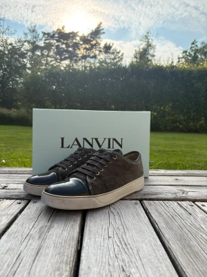 Lanvin svarta låga sneakers - Snygga svarta sneakers från Lanvin med ovandel i mocka och tåparti i blankt läder. Skorna har vita sulor och svarta skosnören. Perfekta för dig som gillar stilrena och exklusiva sneakers med en twist.