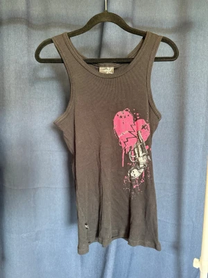 Svart ribbad linnetopp från Darkside - Cool svart ribbad linnetopp från Darkside med tryck av ett rosa hjärta och en revolver på framsidan. Toppen är ärmlös och har en tight passform, perfekt för dig som gillar alternativ stil och rockiga detaljer.