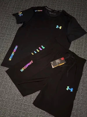 Svart träningsset från Under Armour - Säljer en svart träningsset från Under Armour med färgglad logga och textdetaljer. Shortsen har snörning i midjan och är tillverkade i ett mjukt, syntetiskt material som passar perfekt för träning eller chill. Matchar med t-shirt i samma stil. Storlek S, helt ny 