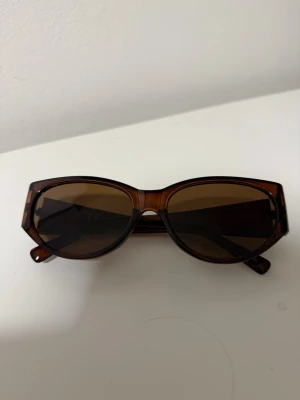 Bruna solglasögon med bred båge - Snygga solglasögon med bred, brun båge och mörka glas. Modellen har en lätt kantig form som ger en cool retrovibe. Perfekta för soliga dagar och passar till de flesta outfits.