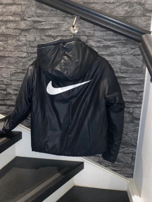 Svart Nike vindjacka med huva - Snygg svart vindjacka från Nike med stor vit logga på ryggen och mindre logga på bröstet. Jackan har dragkedja framtill, två sidofickor och en praktisk huva. Man kan böna den ut och in, två olika design och material som man kan välja mellan.