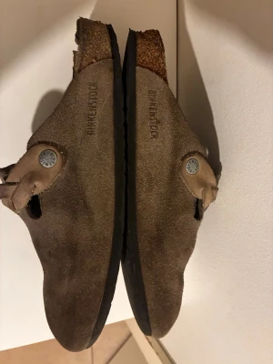 Bruna mockatofflor från Birkenstock - Ett par klassiska bruna tofflor från Birkenstock i mocka med justerbart spänne och korksula. Tofflorna har en stilren design och präglad logga på sidan. Perfekta för dig som gillar en avslappnad och trendig look.