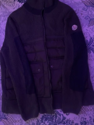 Svart cardiganjacka från Moncler - Snygg svart cardiganjacka från Moncler med stickade ärmar och quiltad front. Jackan har hög krage, dragkedja framtill och två fickor med knappar. Moncler-logga på vänster ärm. Perfekt för lager på lager och streetwear.