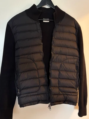 Moncler cardiganjacka - Snygg svart cardiganjacka med quiltad framsida och stickade ärmar samt rygg. Jackan har dragkedja framtill, hög krage och två sidofickor med dragkedja. Perfekt för lager-på-lager och en stilren look.