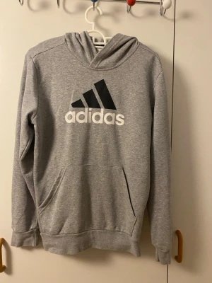 Grå hoodie från Adidas med logga - Snygg grå hoodie från Adidas med stor svartvit logga på bröstet. Klassisk känguruficka framtill och huva. Tillverkad i mjukt sweatshirtmaterial, perfekt för chill eller träning. Ribbade muddar vid ärmslut och nederkant för skön passform.