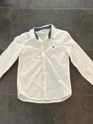Vit skjorta från H&M med kontrastkrage - Klassisk vit skjorta från H&M i bomull med lång ärm och knappar framtill. Skjortan har en snygg kontrastkrage i grått och en liten broderad detalj på bröstet. Perfekt för en clean och stilren look.