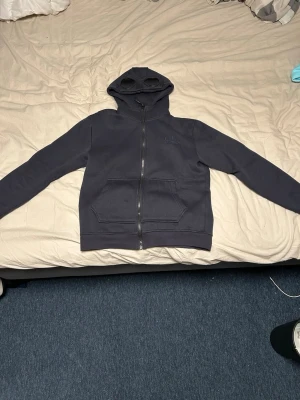 C.P company  - Säljer en mörkblå hoodie från C.P. Company med dragkedja framtill och två stora fickor. Huvan har inbyggda glasögon och coola detaljer med metallknappar. Perfekt för dig som vill sticka ut med streetwear-stil.