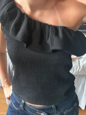 Svart oneshoulder topp med volang - Säljer en svart oneshoulder topp med bred volang längs halsringningen. Toppen är ribbad och har en tight passform som framhäver figuren. Perfekt för dig som vill ha en trendig och stilren look. Har tyvärr ingen lapp men tror att den är gammal från Gina💖💖