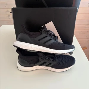 Svarta Adidas Ultraboost sneakers - Säljer ett par svarta Adidas Ultraboost sneakers med vit sula och klassiska tre ränder på sidan. Ovandelen är i ventilerande mesh och skon har en bekväm, responsiv Boost-mellansula. Perfekta för dig som gillar stilrena och sportiga skor.