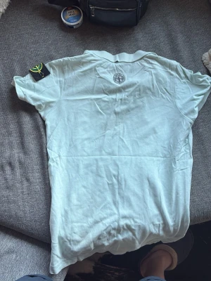 Ljusgrön pikétröja från Stone Island - Säljer en vit pikétröja från Stone Island med klassisk krage och knappar framtill. Tröjan har korta ärmar och den ikoniska Stone Island-loggan på bröstet samt patch på ärmen. Perfekt för en clean och stilren look.