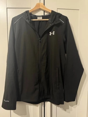 Svart vindjacka från Under Armour - Svart vindjacka från Under Armour med huva och hel dragkedja. Jackan har två sidofickor, reflekterande detaljer och logga på bröstet. Är i jätte bra skick som ny och den är i Storlek L men passar även M alltså L/M 