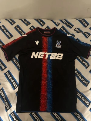 Crystal Palace T-Shirt - Snygg svart matchtröja från Crystal Palace med röd och blå vertikal rand i mitten. Tröjan har klubbmärke på bröstet, sponsorlogga 'NET88' och röda detaljer vid krage och ärmslut. Perfekt för dig som älskar fotboll och vill sticka ut på planen.