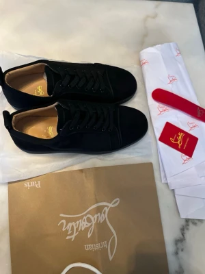 Svarta Christian Louboutins - Stilrena svarta sneakers från Christian Louboutin med klassisk röd sula och diskret logga på insidan. Skorna har en låg profil, svarta skosnören och ovandel i mocka. Perfekta för dig som vill ha en lyxig touch på din outfit.