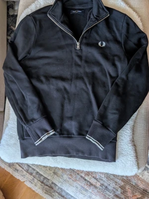 Svart halfzip tröja från Fred Perry - Svart långärmad tröja från Fred Perry med halv dragkedja och broderad logga på bröstet. Tröjan har ribbade muddar och nederkant med vita ränder för en sportig look. Perfekt för en stilren och avslappnad outfit. Den är nästan helt ny har använt en gång 