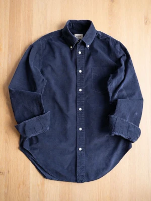 Mörkblå manchesterskjorta från Bellerose - Säljer en mörkblå manchesterskjorta från Bellerose med klassisk button-down krage och vita knappar. Skjortan har en bröstficka och lång ärm, perfekt för en avslappnad men ändå stilren look.